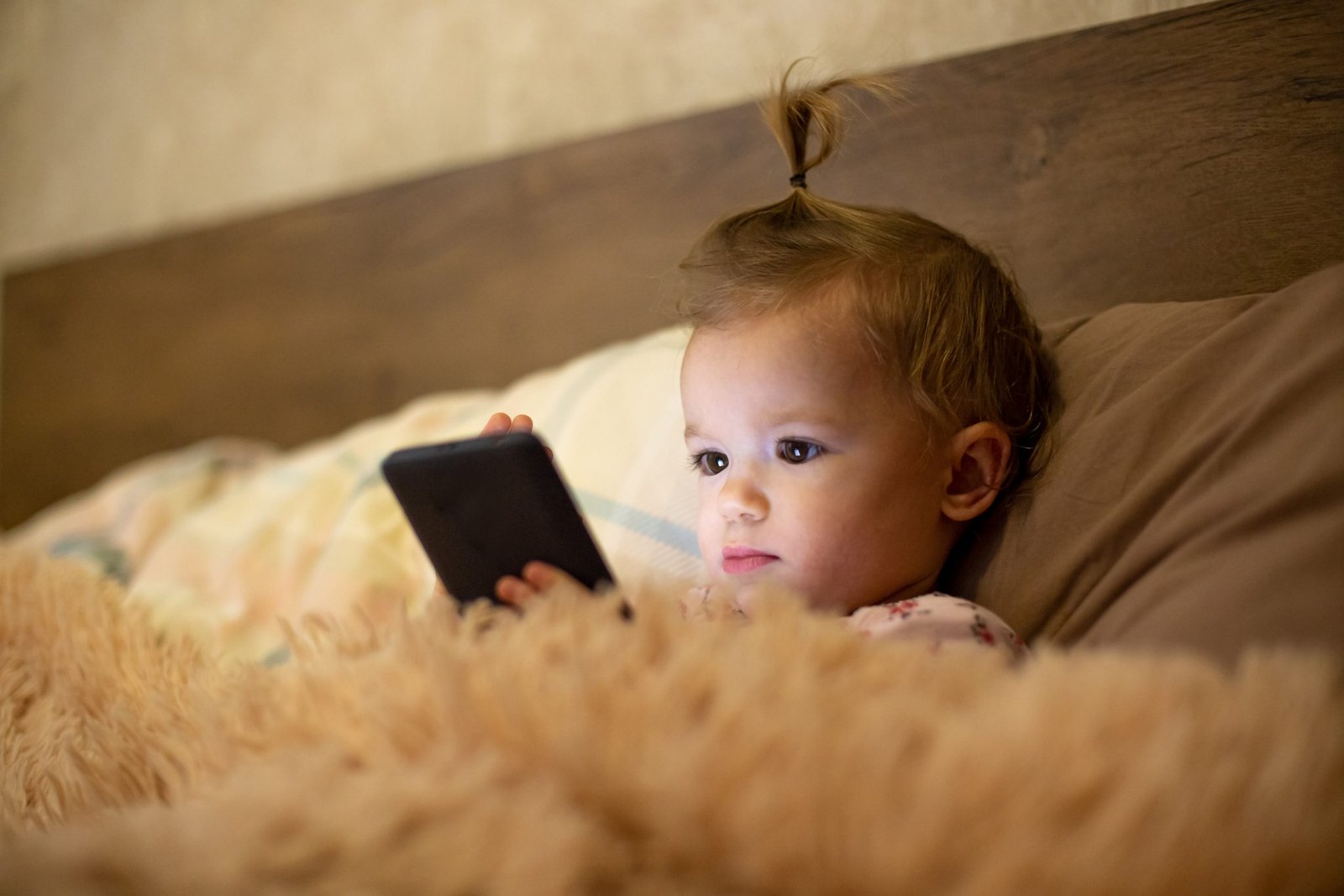 a little girl lying in bed in a room with the light off and holding a smartphone, playing games and watching cartoons. Baby addiction to gadgets